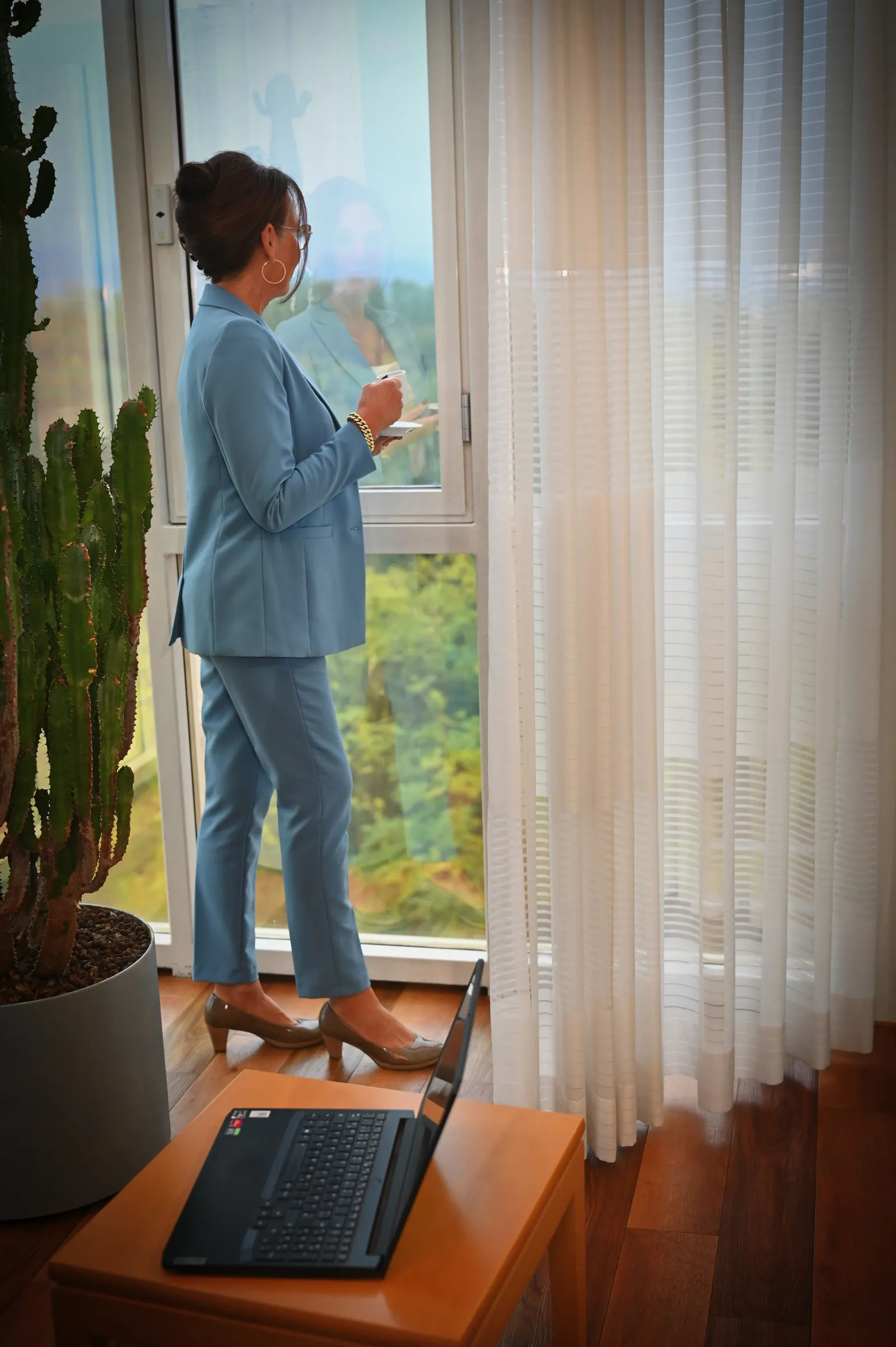 Sabine Heyne steht im blauen Anzug mit einer Tasse am Fenster, im Vordergrund ein Laptop und ein Kaktus